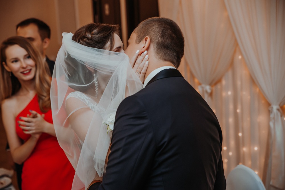 Pocałunek nowożeńców: panna młoda w welonie i sukni ślubnej, pan młody w czarnym garniturze, w tle goście weselni i dekoracja z jasnych zasłon z lampkami.