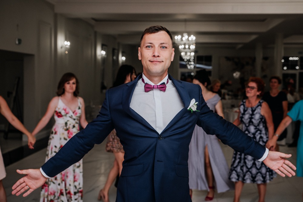 Uśmiechnięty mężczyzna w granatowym garniturze i bordowej muszce, z rozłożonymi ramionami, otoczony przez tańczących gości weselnych trzymających się za ręce w jasnej sali balowej z żyrandolami.