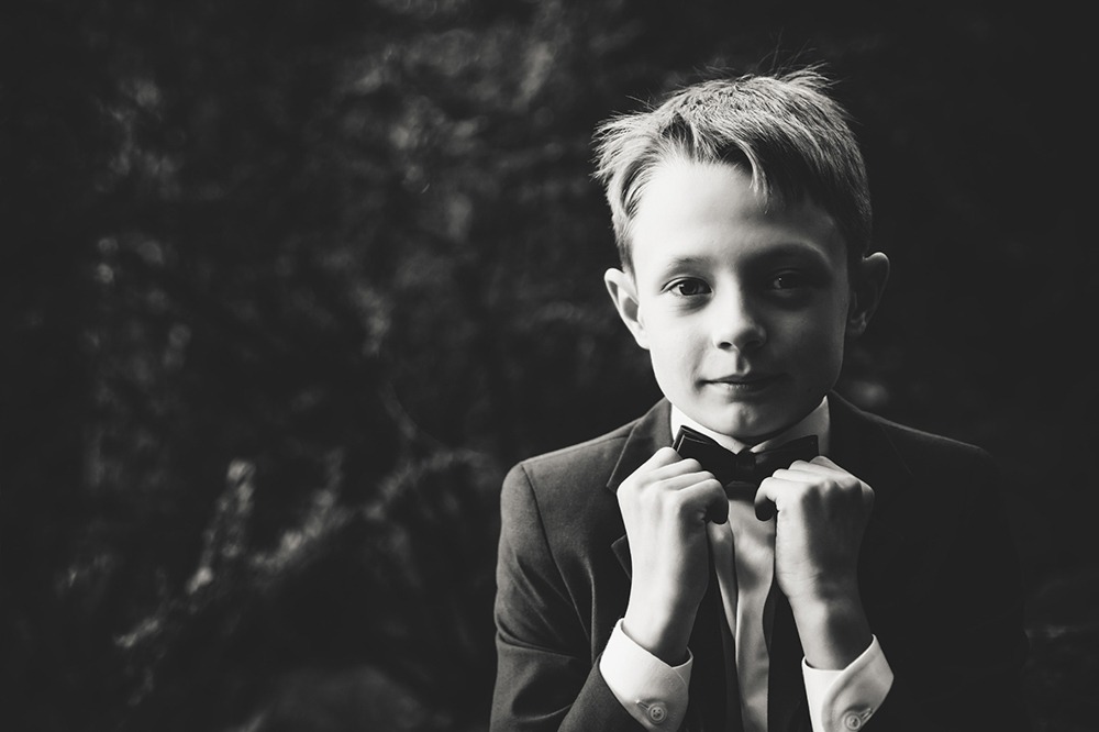 Czarno-białe portretowe ujęcie młodego chłopca w garniturze z muszką, poprawiającego ją dłońmi, na rozmytym ciemnym tle.