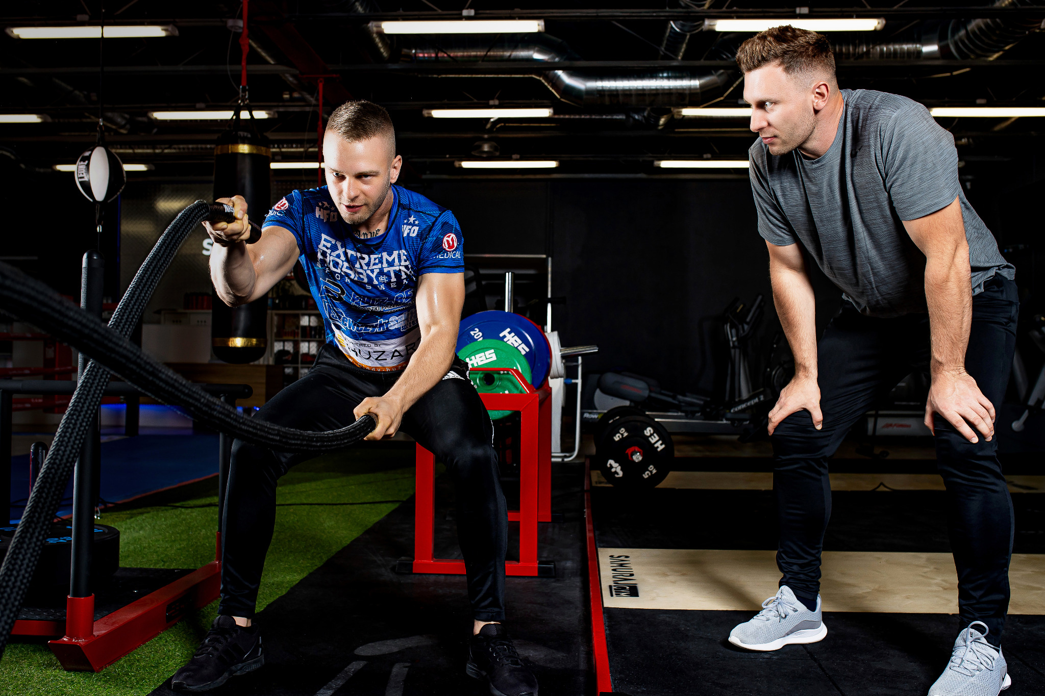 Mężczyzna w sportowej koszulce ćwiczy z grubymi linami pod okiem trenera w siłowni, widoczne worki bokserskie i sprzęt do podnoszenia ciężarów.