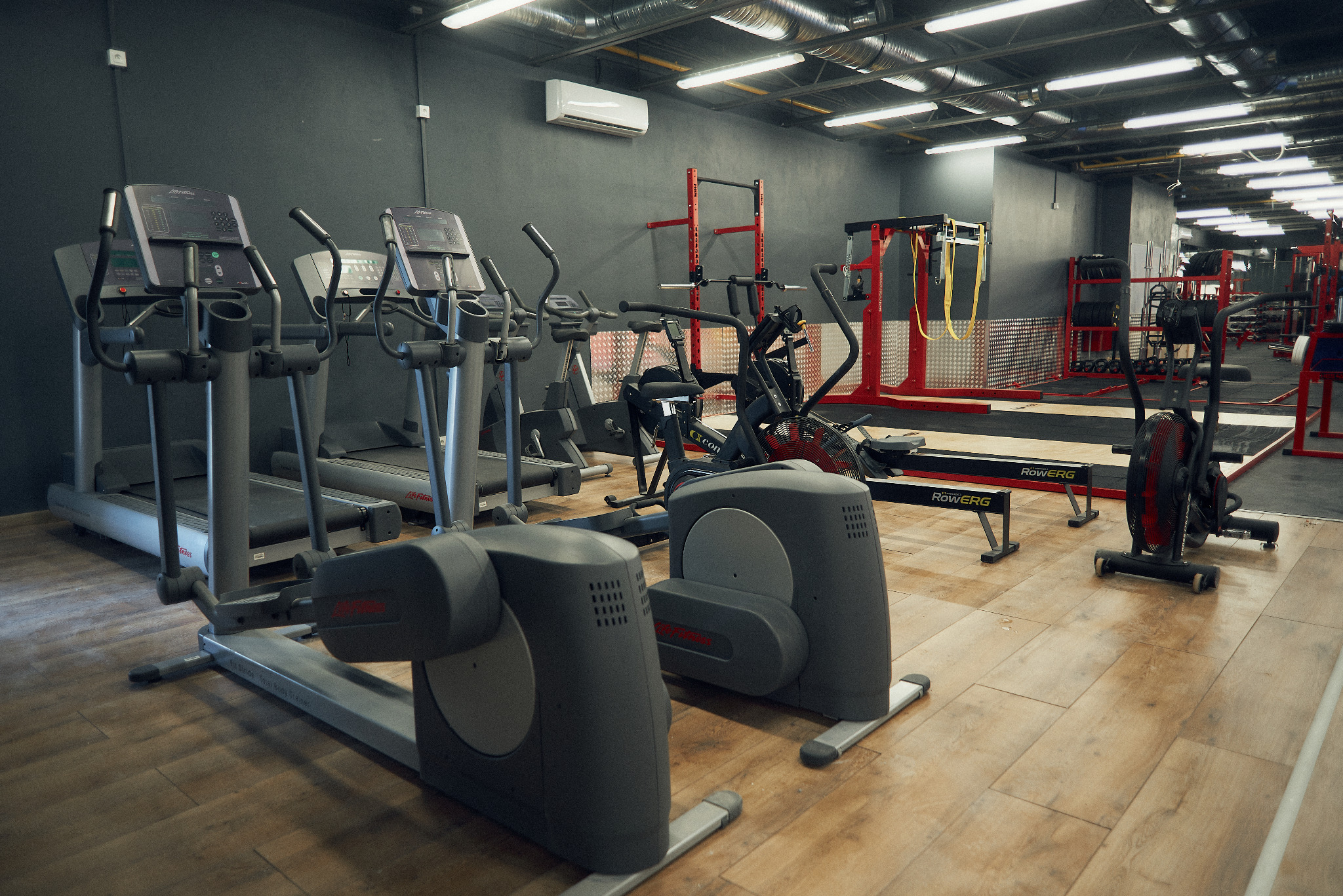 Widok na salę fitness z różnorodnym sprzętem: bieżnie, orbitreki, rowerki powietrzne i wioślarze, ustawione na drewnianej podłodze, z szarą ścianą w tle i systemem oświetlenia sufitowego.