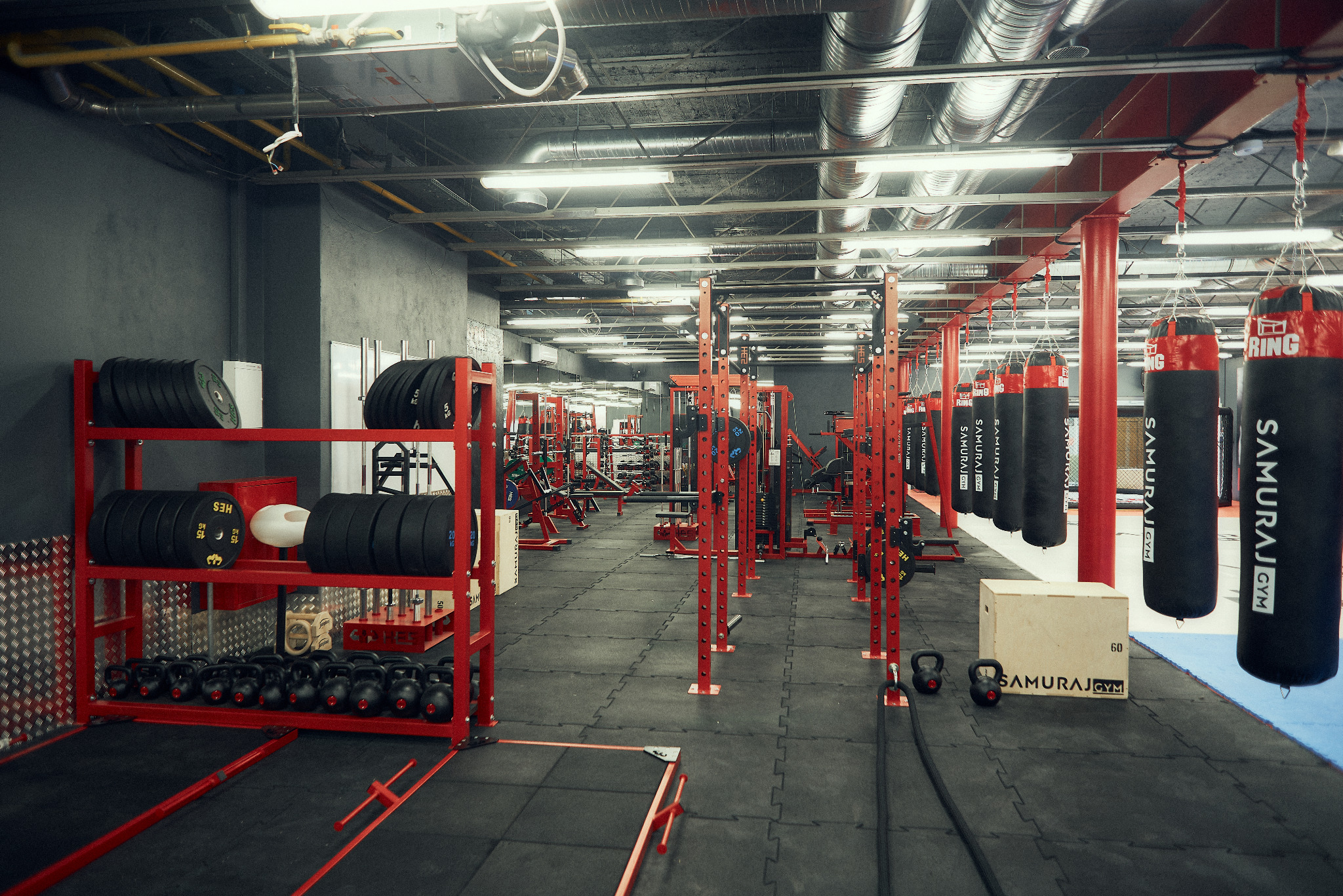 Wnętrze siłowni z czerwonymi konstrukcjami do ćwiczeń, rzędem czarnych worków bokserskich z napisem SAMURAJ GYM, stojakiem z obciążeniami i kettlebells, oraz gumową podłogą.