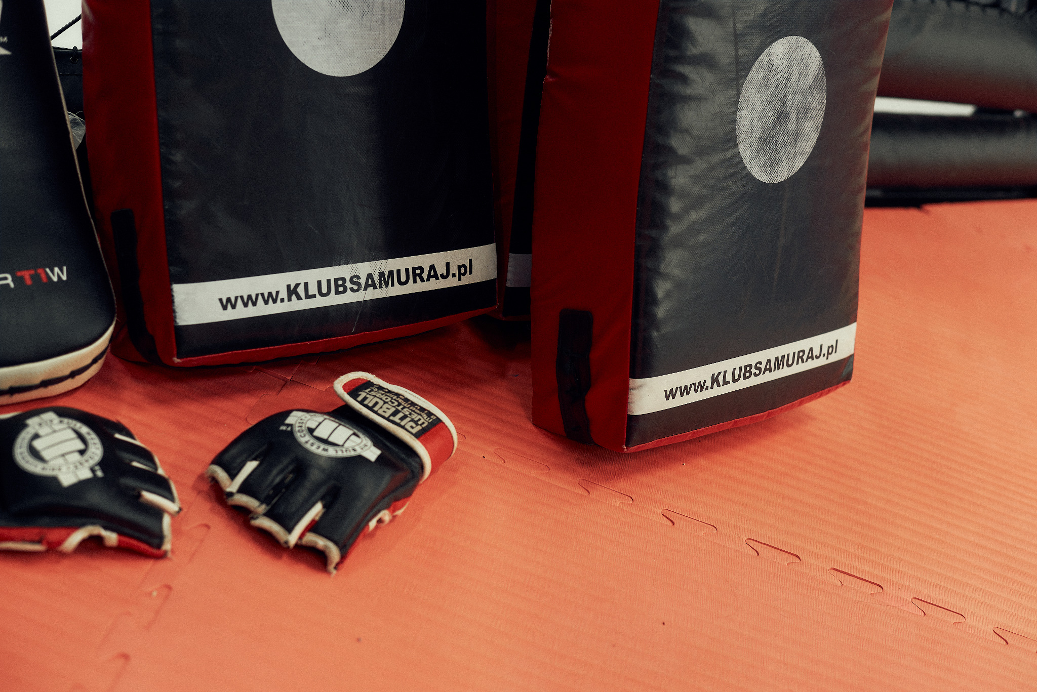 Sprzęt do sportów walki: tarcze z logo www.KLUBSAMURAJ.pl oraz rękawice MMA Pit Bull, leżące na pomarańczowej macie treningowej.