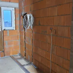 Wewnętrzne ściany z czerwonej cegły z bruzdami, w kt&oacute;rych poprowadzone są białe kable elektryczne. Na podłodze widoczna metalowa konstrukcja pod podłogę techniczną i narzędzia.