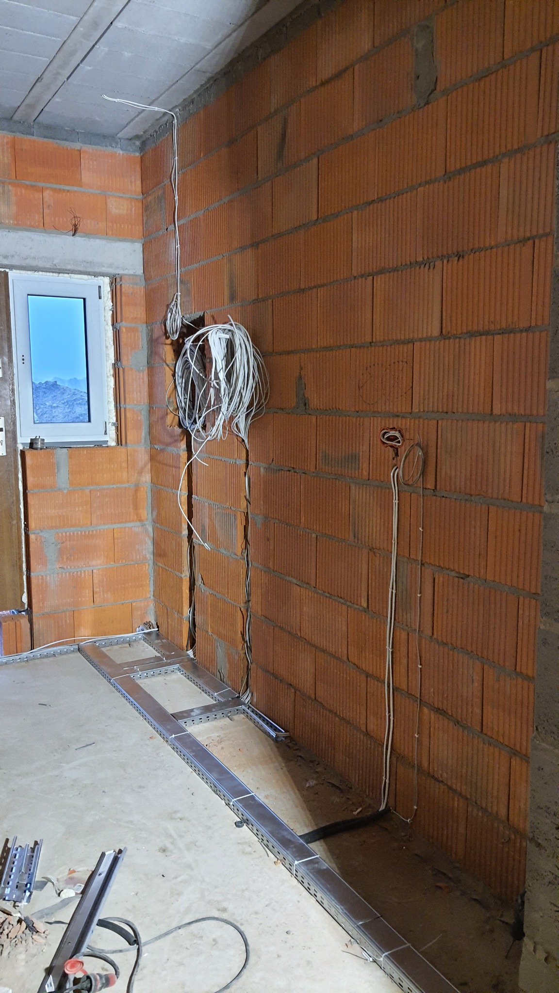 Wewnętrzne ściany z czerwonej cegły z bruzdami, w których poprowadzone są białe kable elektryczne. Na podłodze widoczna metalowa konstrukcja pod podłogę techniczną i narzędzia.