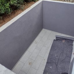 Firma Cyprian - Świeżo pomalowany na szaro wnęka z wyłożoną płytkami podłogą, zabezpieczoną szarą włókniną malarską z rolką taśmy malarskiej, widok z góry na zewnątrz z zielonymi krzewami w tle.