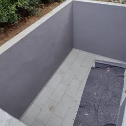 Świeżo pomalowany na szaro wnęka z wyłożoną płytkami podłogą, zabezpieczoną szarą włókniną malarską z rolką taśmy malarskiej, widok z góry na zewnątrz z zielonymi krzewami w tle.