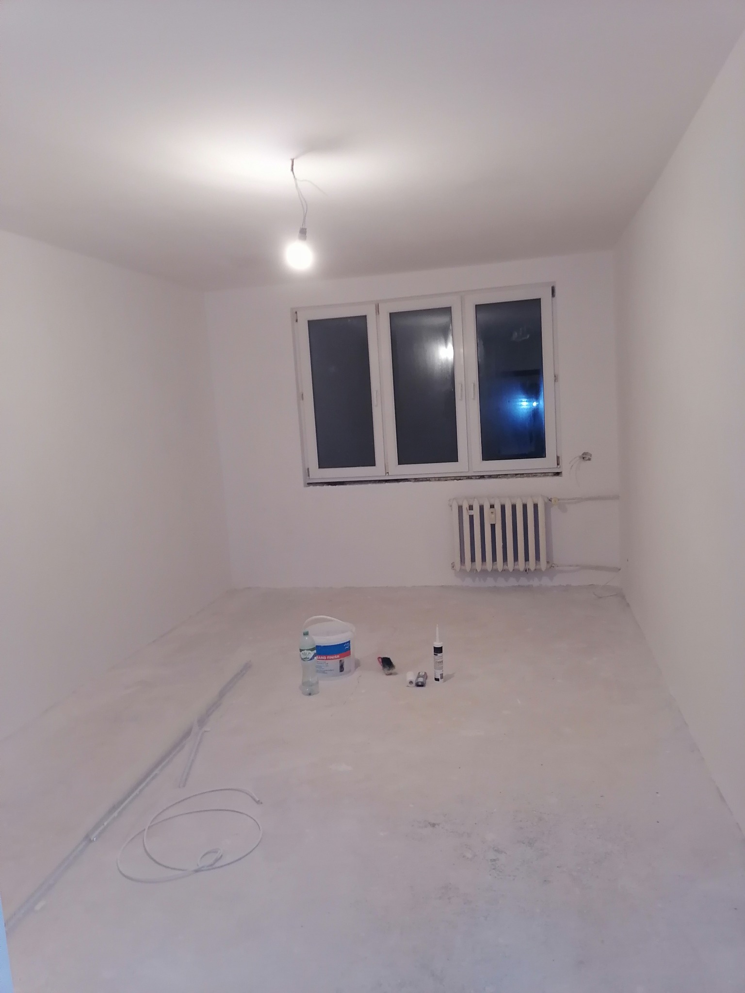 Wnętrze pokoju w trakcie prac przygotowawczych: białe ściany, zakurzona podłoga, grzejnik pod oknem, wiadro z farbą i akcesoria malarskie na podłodze, kabel zwisający z sufitu.