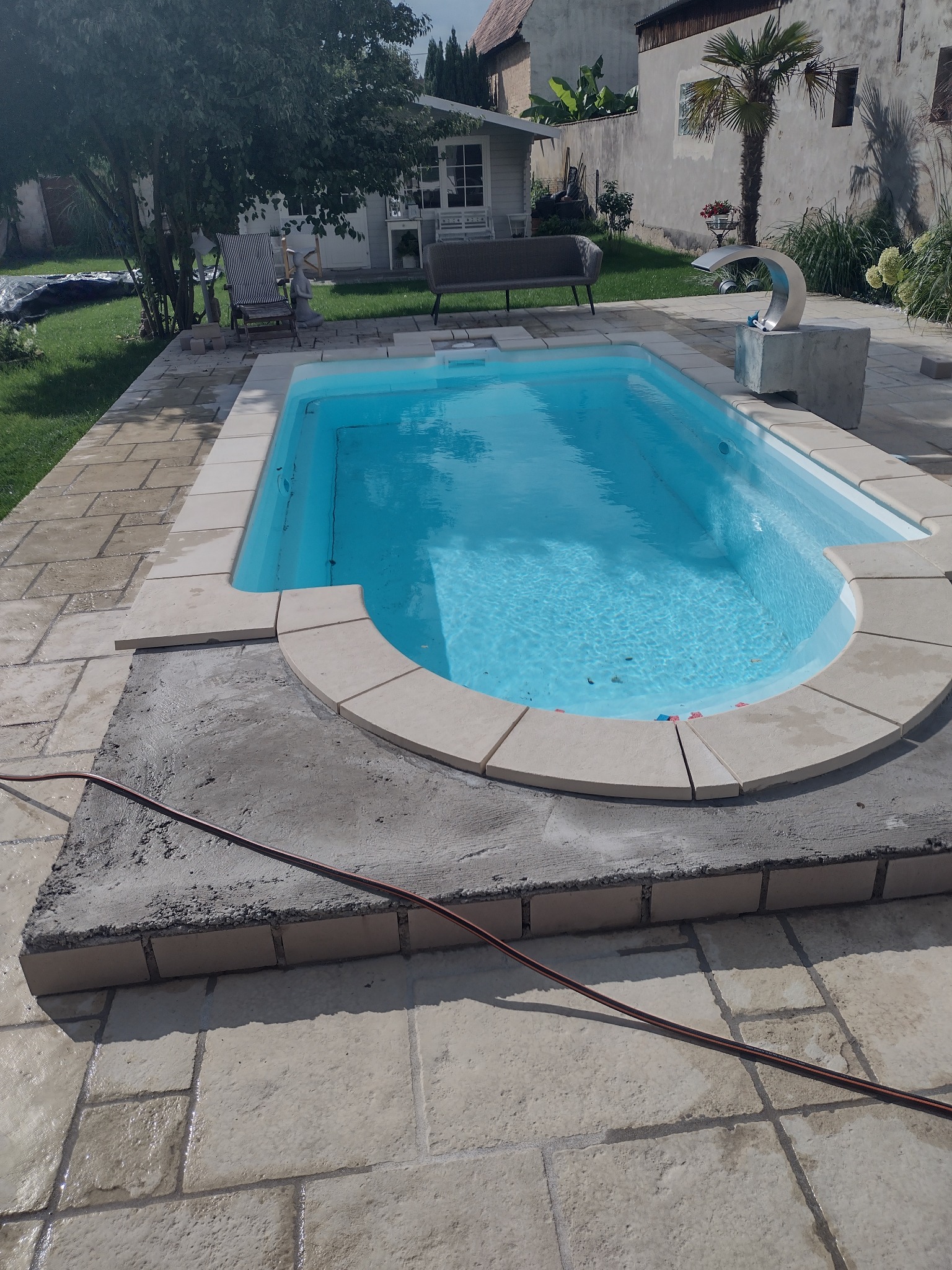 Niebieski basen ogrodowy z obudową z jasnych płytek, częściowo wykończony, widoczny wąż ogrodowy na posadzce z kamiennych płyt, w tle trawnik, drzewa i zabudowania.