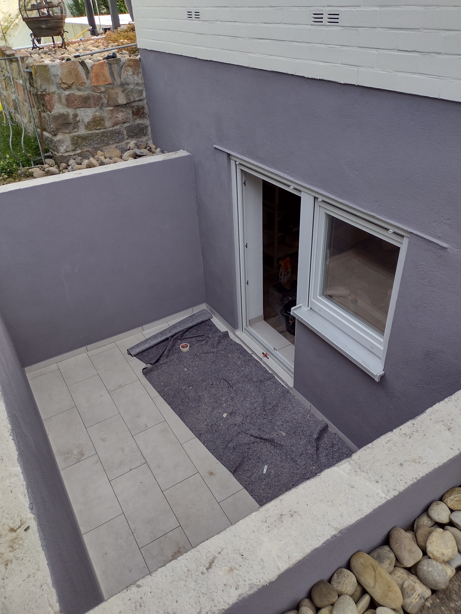 Wykończona wnęka z szarą ścianą, białymi płytkami podłogowymi i oknem, zabezpieczona szarą włókniną malarską, widok z góry.