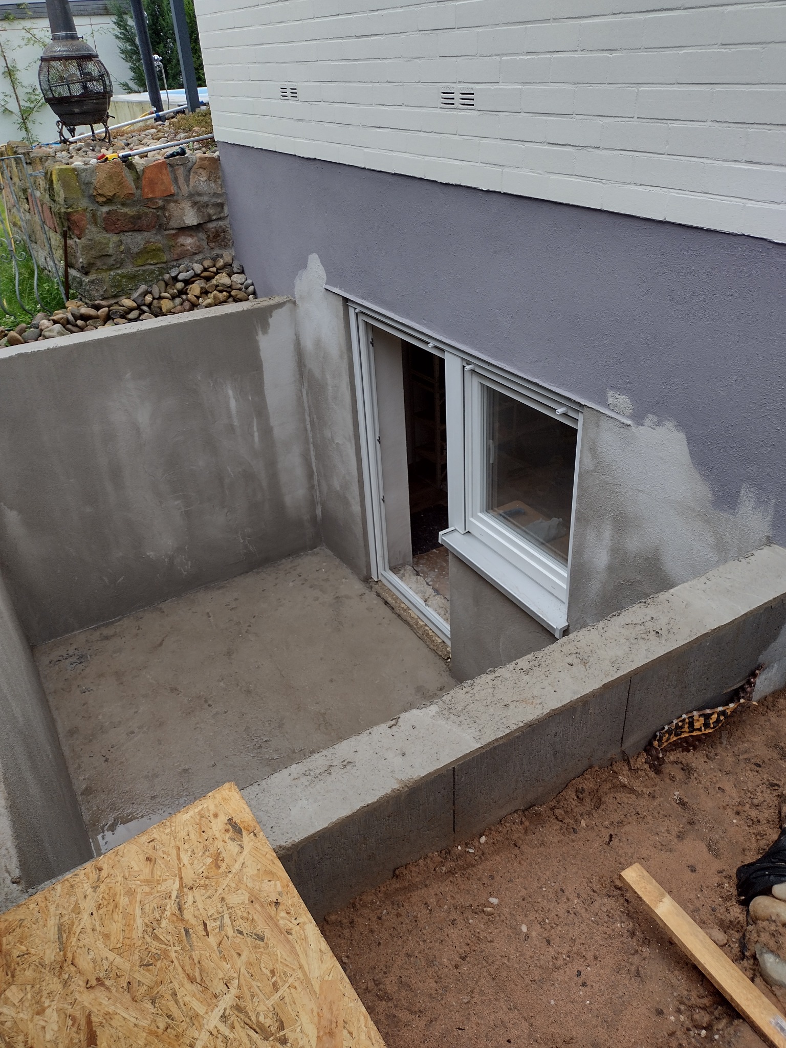 Wybetonowane zejście do piwnicy z nową, białą ramą okienną, fragment ściany zewnętrznej pomalowany na szaro, widoczne ślady świeżego betonu i drewniana płyta na ziemi.