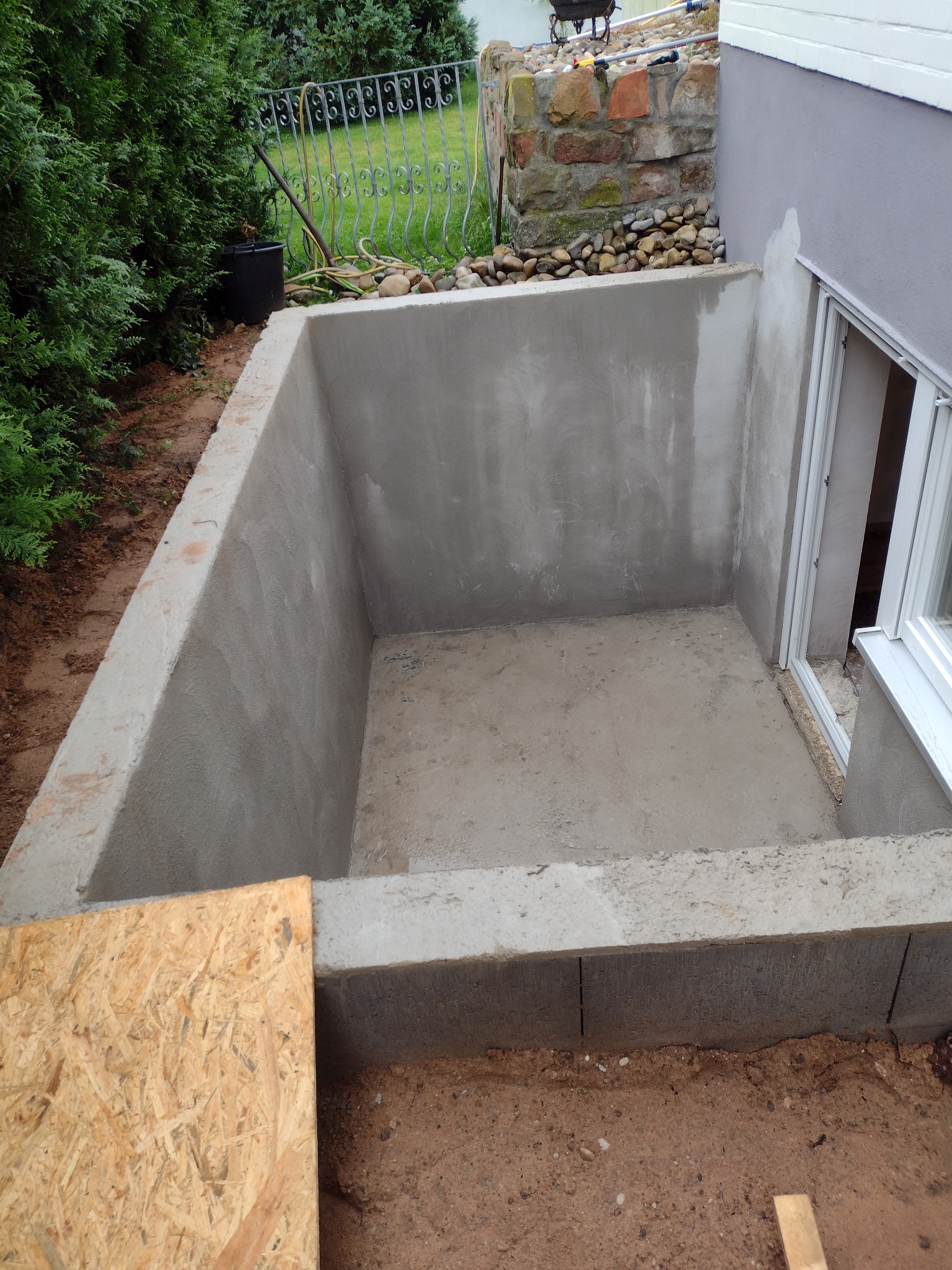 Betonowa piwnica zewnętrzna z oknem po remoncie, widoczna surowa płyta OSB, krzewy w tle.