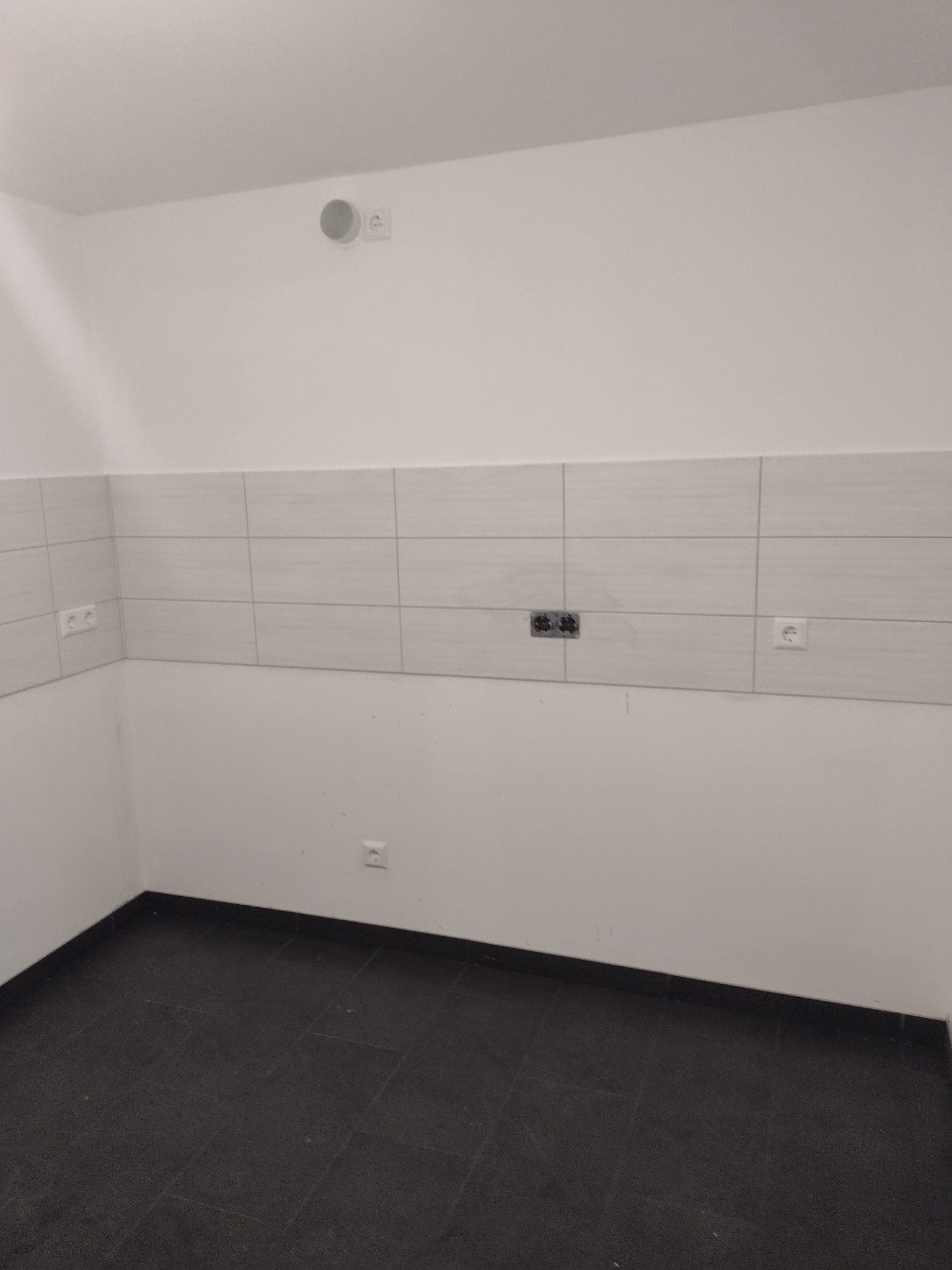 Pusta kuchnia w trakcie prac wykończeniowych: białe ściany, szare płytki nad blatem, czarna podłoga z płytek, widoczne gniazdka elektryczne i otwór na instalację.