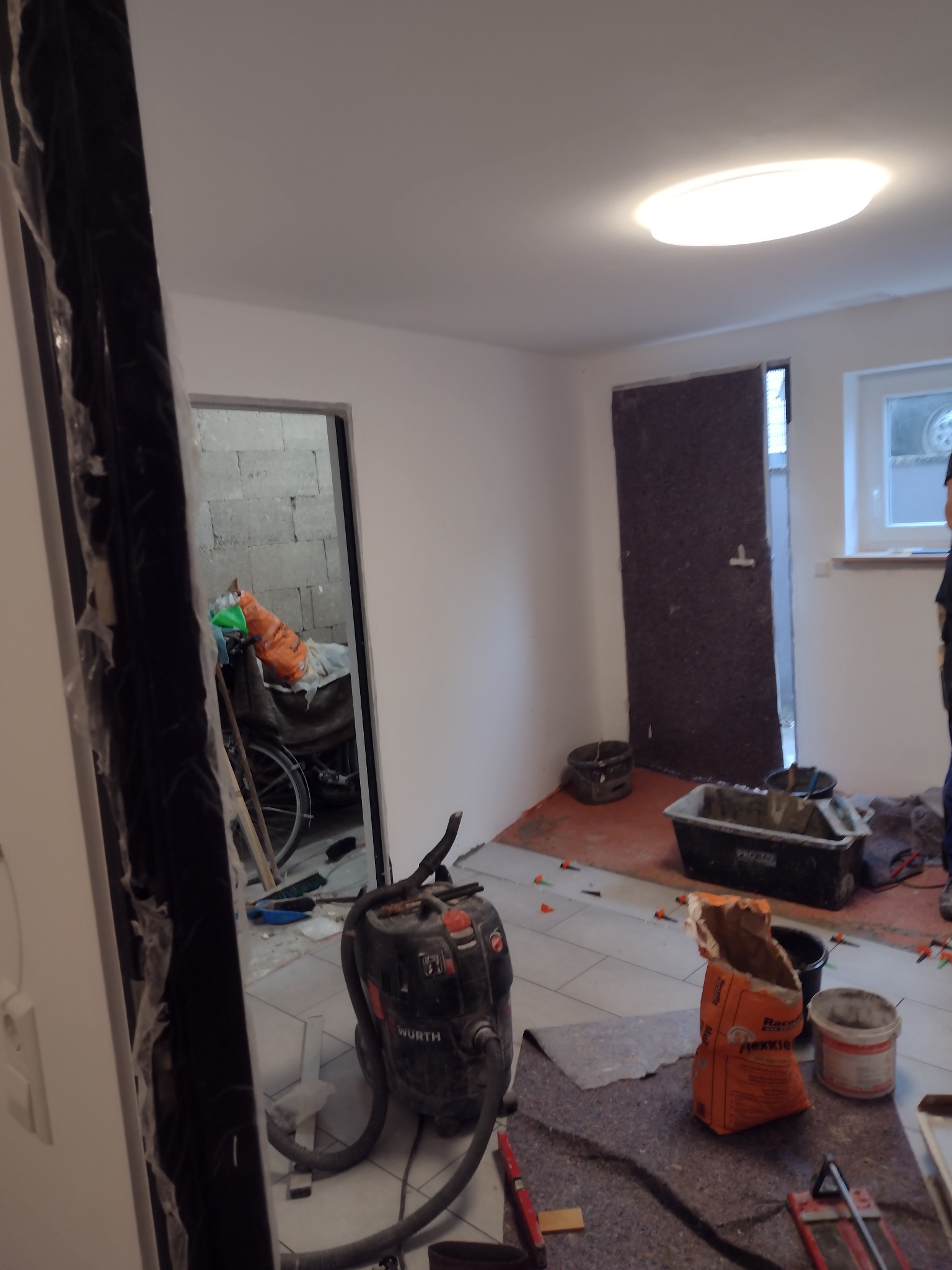 Wnętrze w trakcie prac, widoczny odkurzacz przemysłowy Wurth, worek z klejem Flexkleber, wiadro, narzędzia do układania płytek oraz fragment ułożonej podłogi z płytek ceramicznych.