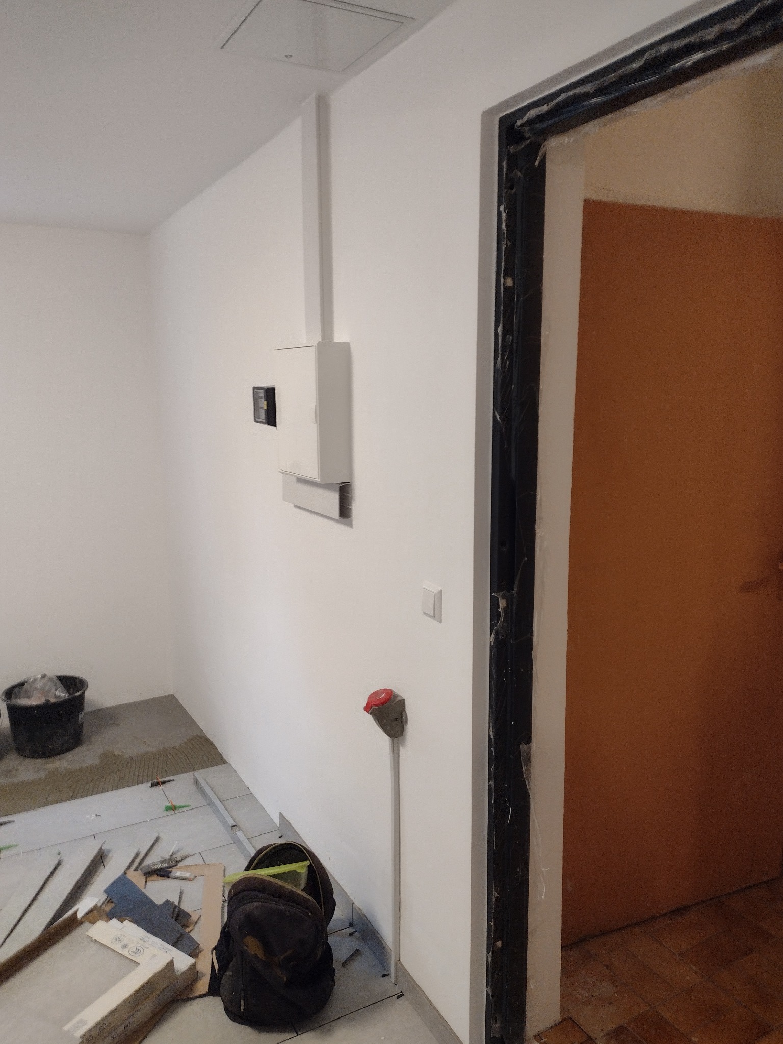 Wnętrze w trakcie prac: białe ściany, osprzęt elektryczny, zabezpieczona futryna drzwi, narzędzia i materiały budowlane na podłodze z ułożonymi płytkami.