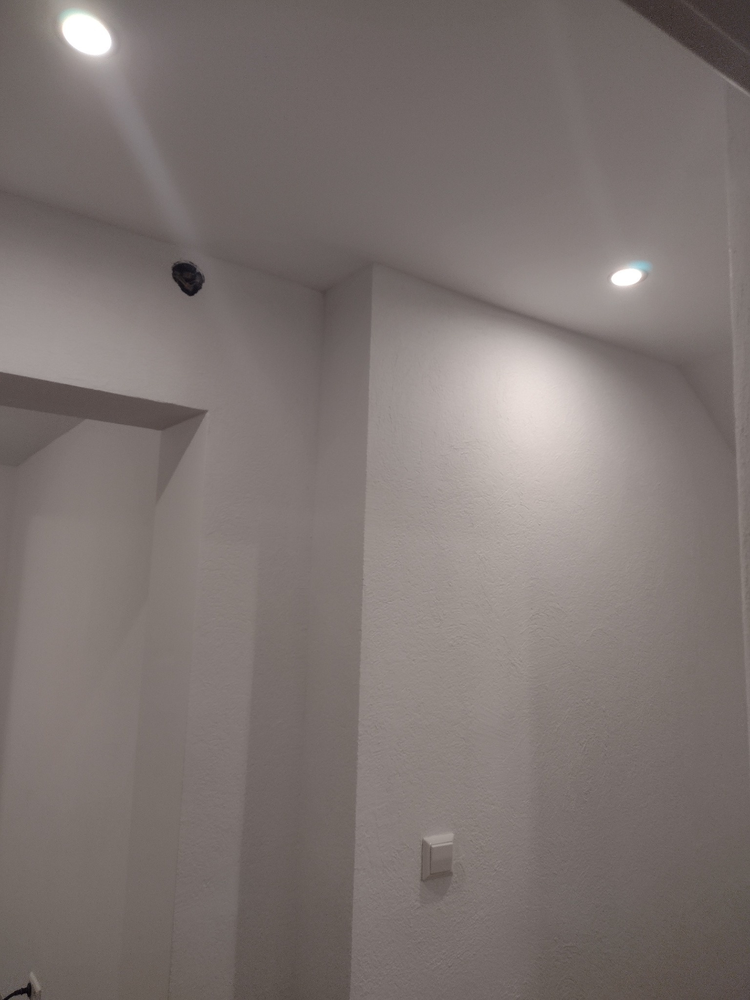 Fragment wnętrza po odświeżeniu: białe ściany z widocznym włącznikiem światła, dwa punkty oświetleniowe w suficie, jeden z nich jeszcze bez oprawy, widoczny otwór montażowy.