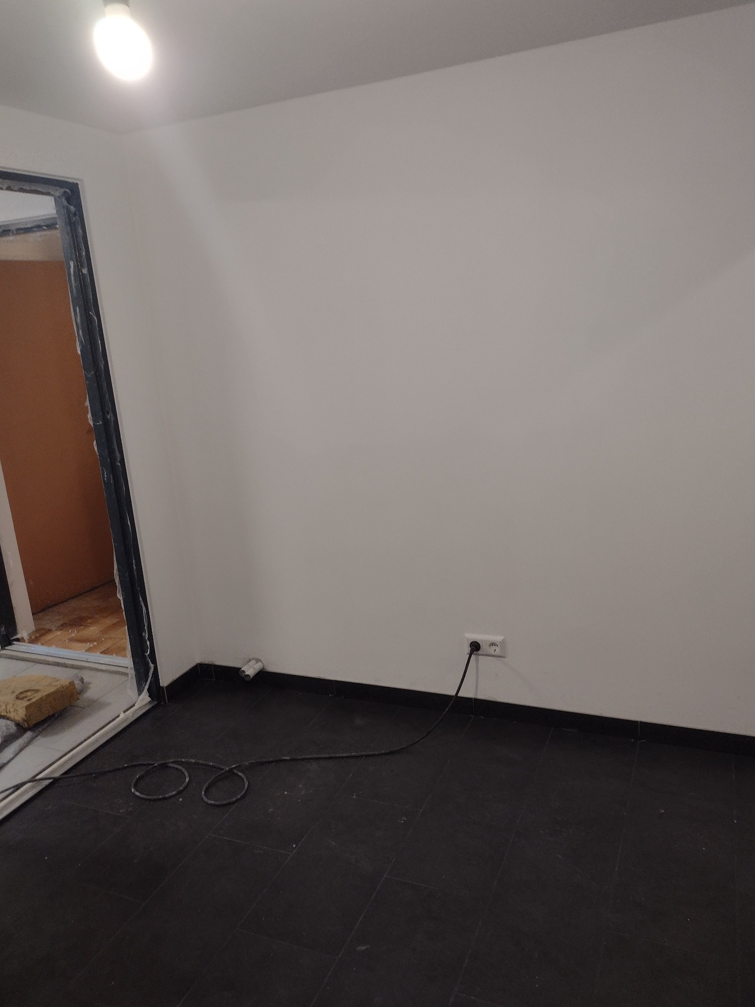 Pusta ściana po malowaniu na biało z czarną podłogą, widoczne gniazdko elektryczne i kabel oraz ościeżnica drzwi z niedokończonym wykończeniem.