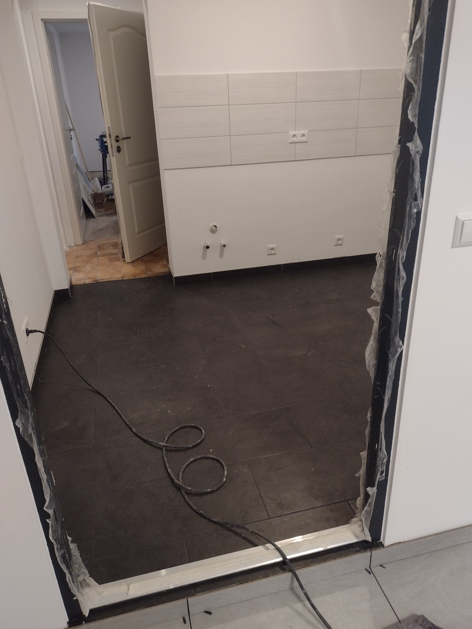 Ciemnoszara podłoga z płytek w trakcie układania, widoczne kable elektryczne, zabezpieczona folią ościeżnica drzwiowa i fragment białej ściany z płytkami oraz gniazdkami elektrycznymi w tle.