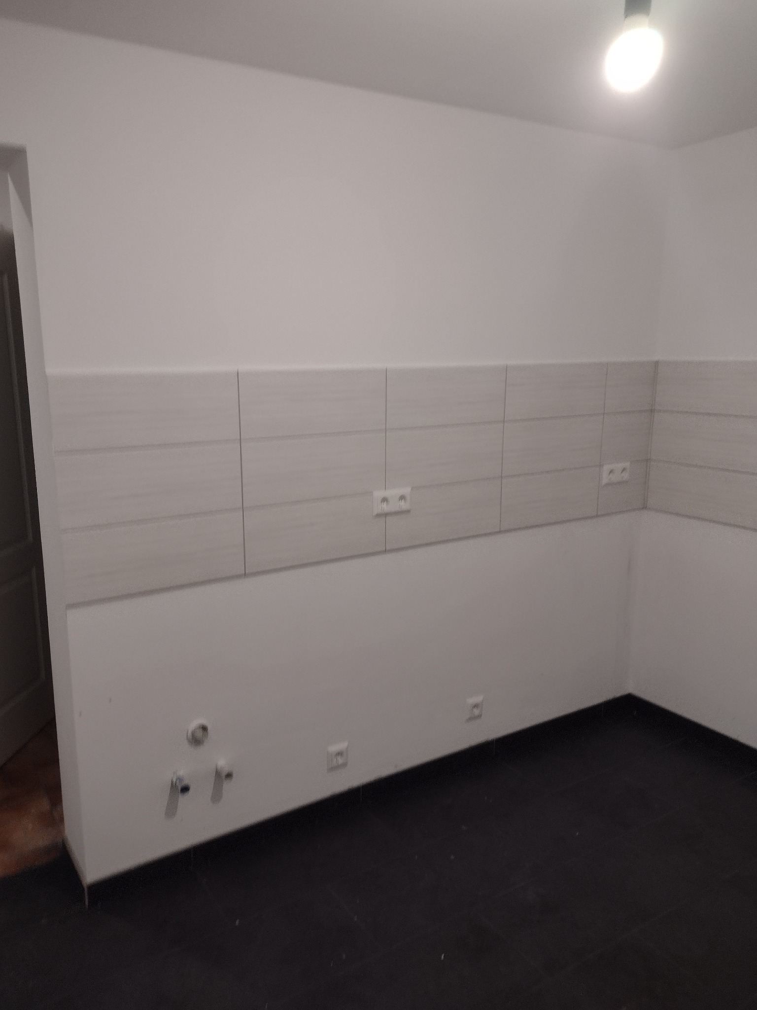 Świeżo wykafelkowana ściana w kuchni z widocznymi gniazdkami elektrycznymi i wyprowadzeniami hydraulicznymi, czarna podłoga, surowy stan przed montażem mebli.