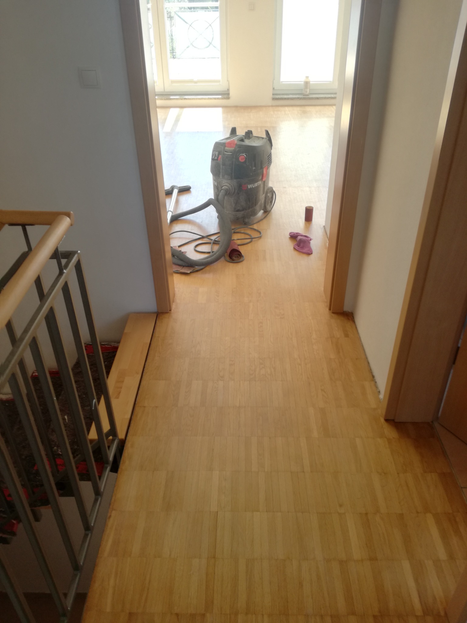 Świeżo położony parkiet w korytarzu, widoczny odkurzacz przemysłowy Wurth, pozostawione różowe kapcie i niedopita puszka po napoju.