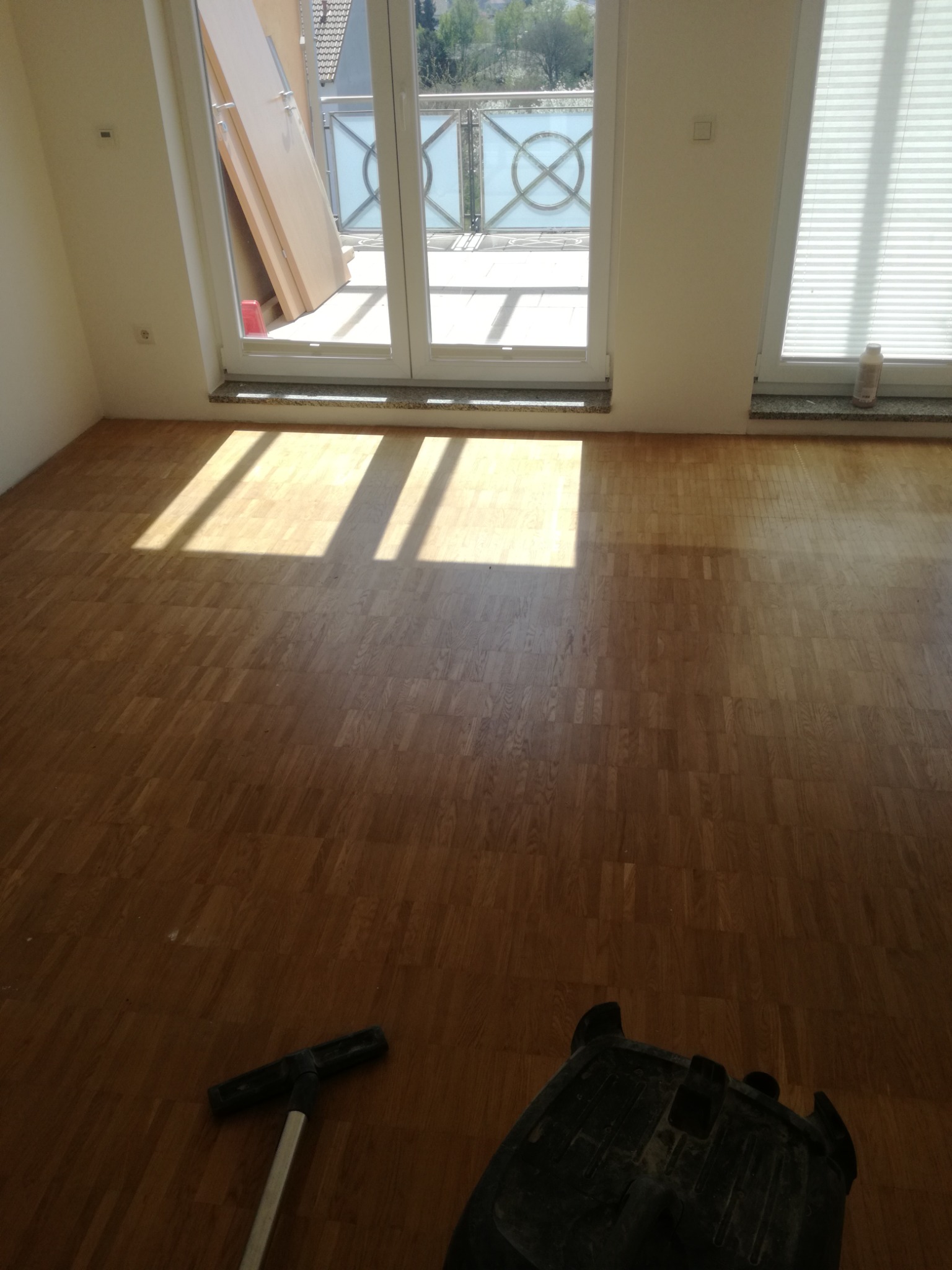 Pokój z nowo ułożonym parkietem, widoczne promienie słoneczne wpadające przez balkonowe okno oraz fragment maszyny do polerowania podłóg.