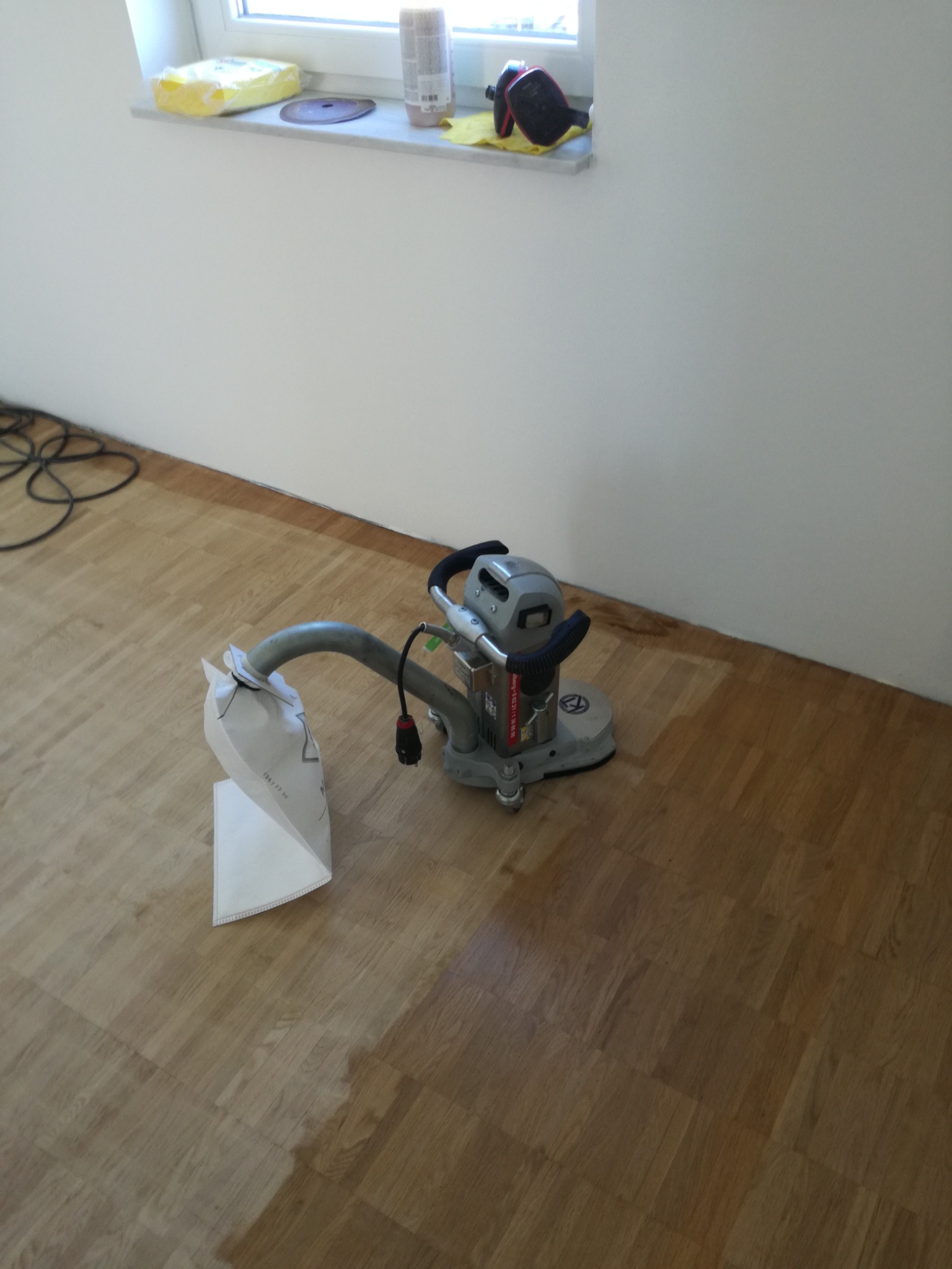 Szlifowanie parkietu maszyną z odsysaniem pyłu, widoczny fragment wyszlifowanej powierzchni, na parapecie okna akcesoria i materiały eksploatacyjne.