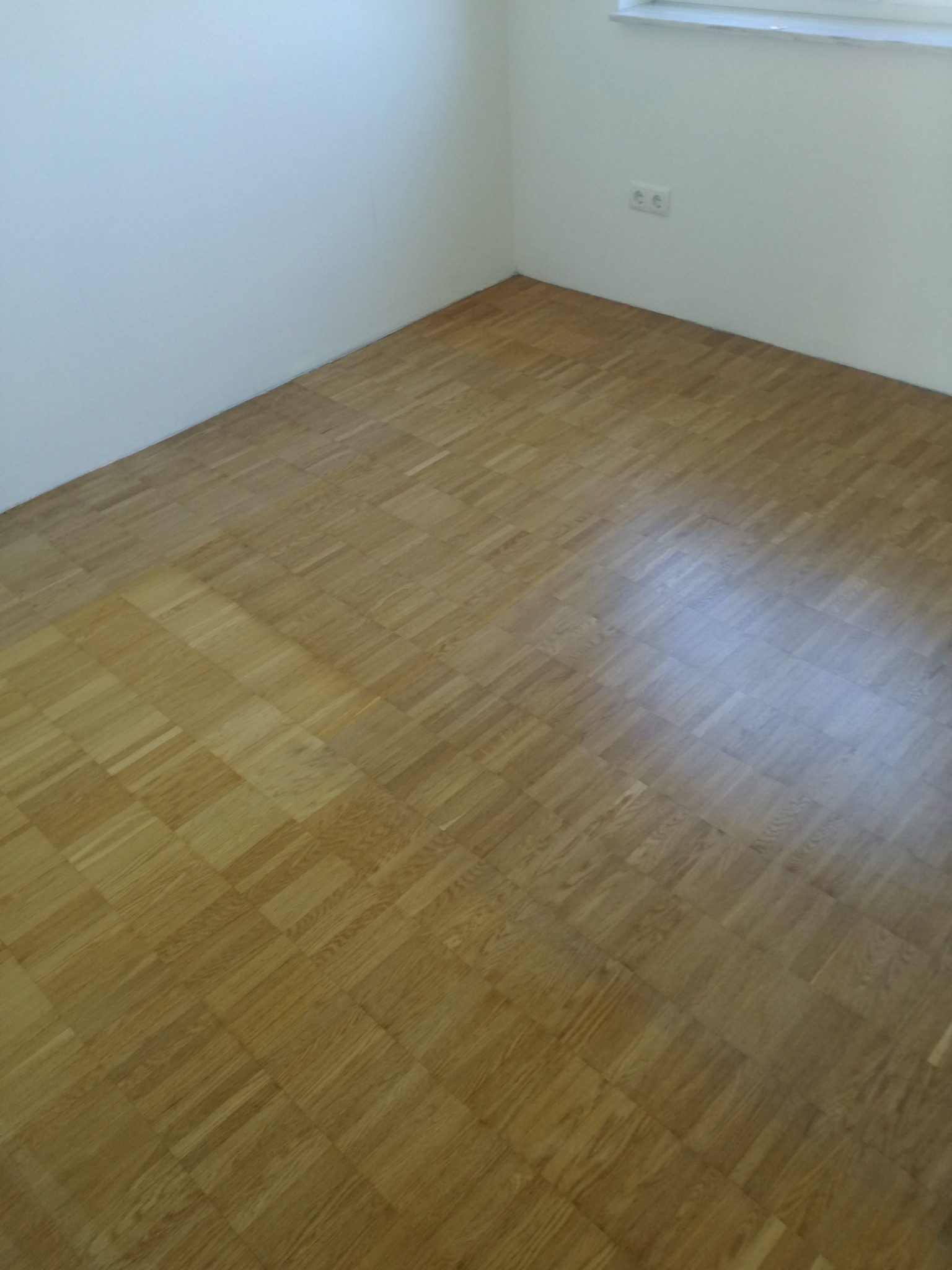 Parkiet ułożony w jodełkę kwadratową, widoczne gniazdka elektryczne przy oknie, białe ściany w rogu pokoju.