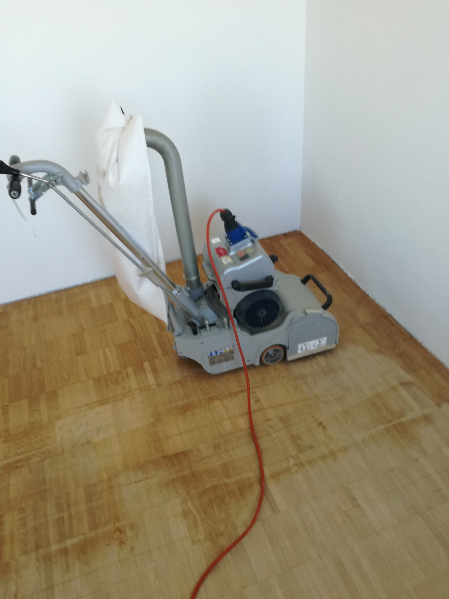 Szlifowanie parkietu maszyną z workiem na pył w rogu pomieszczenia, widoczny czerwony kabel zasilający.