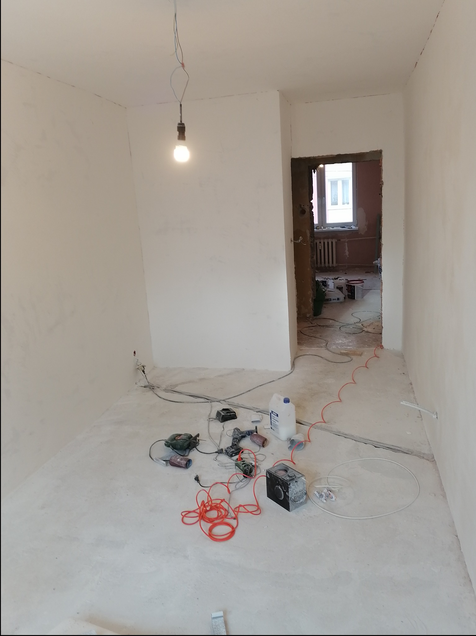 Pokój w trakcie prac wykończeniowych: ściany pokryte gładzią, wisząca żarówka, narzędzia i materiały budowlane na podłodze, widok na sąsiednie pomieszczenie w trakcie remontu.