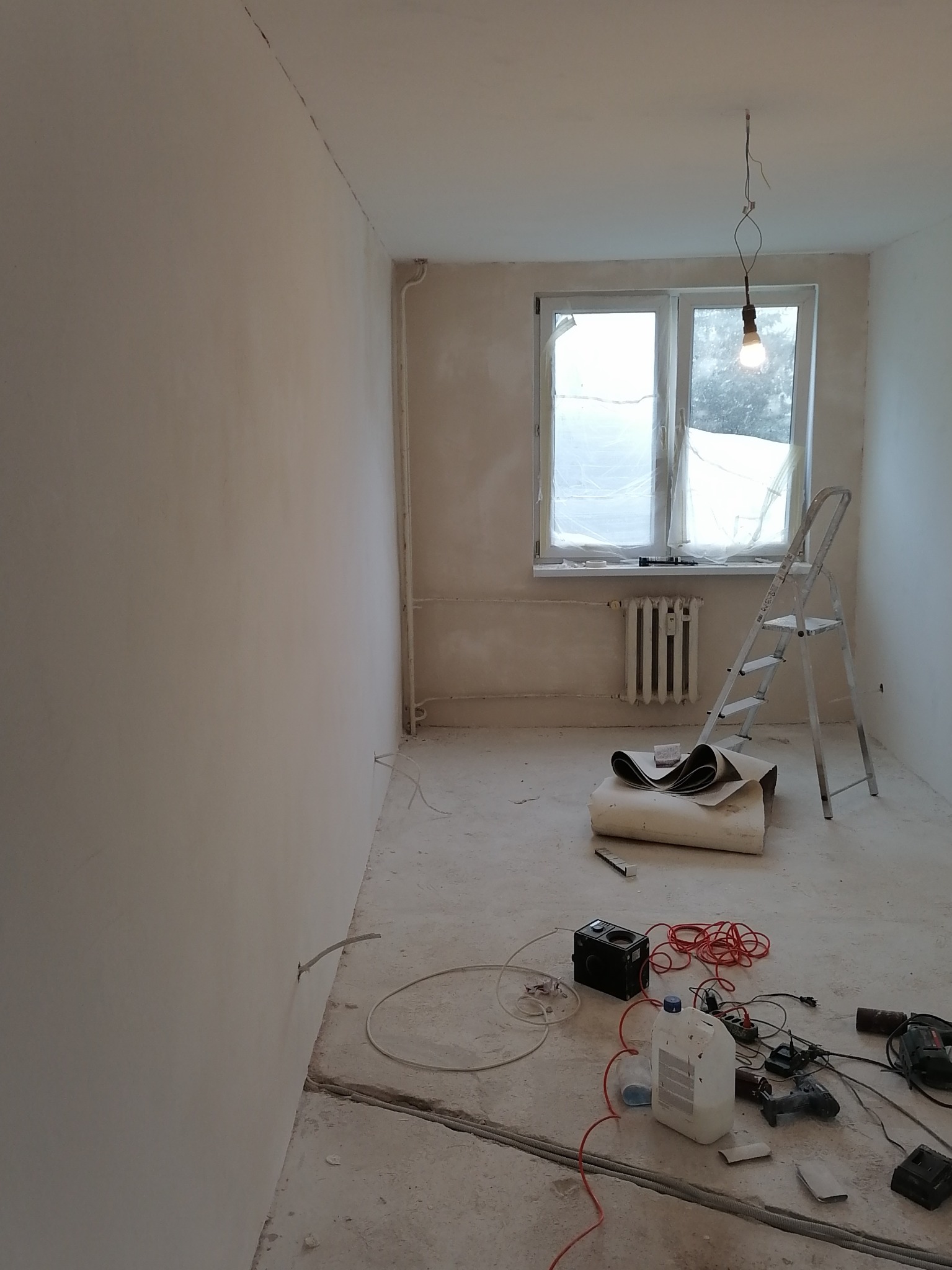 Pokój w trakcie prac przygotowawczych: ściany po nałożeniu gładzi, okno zabezpieczone folią, drabina oparta o ścianę, na podłodze rozłożone narzędzia i materiały.
