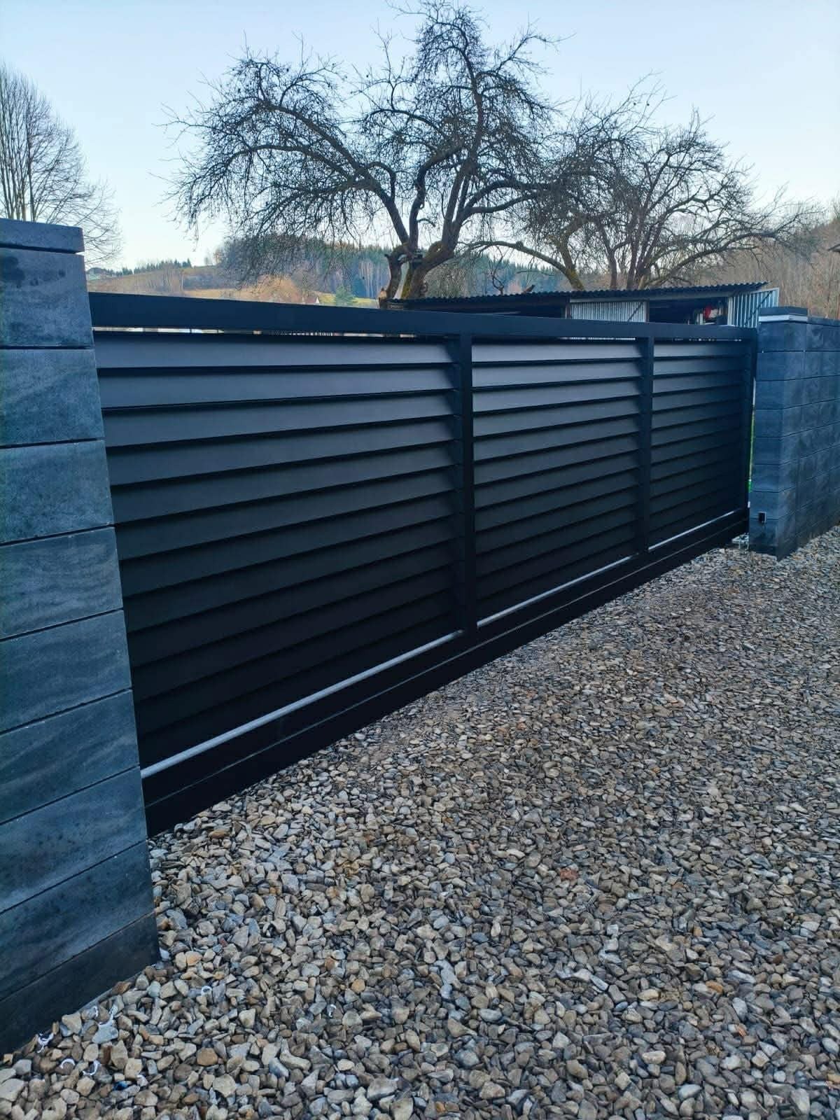 Nowoczesna, przesuwna brama lamelowa w kolorze antracytowym, zamontowana na posesji z wysypanym kamieniem ozdobnym. Widoczne słupki z szarej kostki.