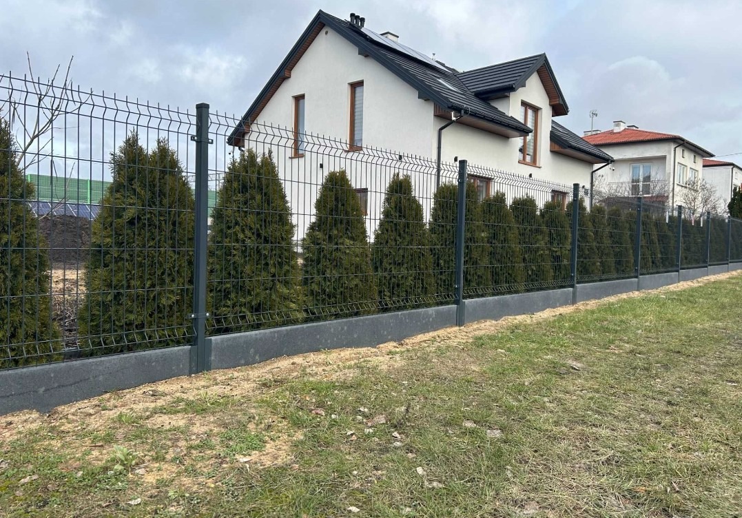 Nowoczesne ogrodzenie panelowe w kolorze grafitowym, z podmurówką i nasadzeniami z tuj, na tle domu z białymi ścianami i ciemnym dachem. Widok z poziomu gruntu.