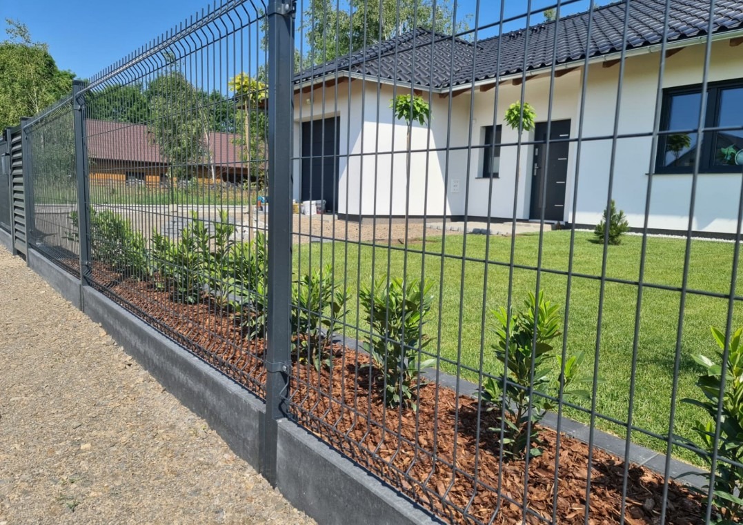 Nowoczesne ogrodzenie panelowe w kolorze grafitowym, z podmurówką i nasadzeniami z roślin, na tle białego domu z czarnym dachem i zadbanym trawnikiem.