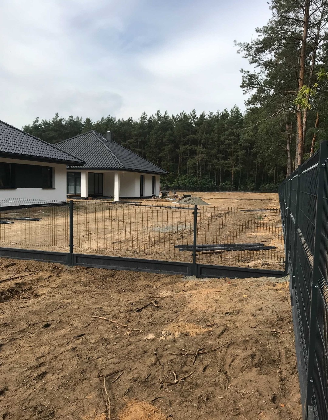 Nowoczesny dom jednorodzinny z czarnym dachem i białymi ścianami, otoczony metalowym ogrodzeniem panelowym na nierównym terenie. W tle widoczny las.