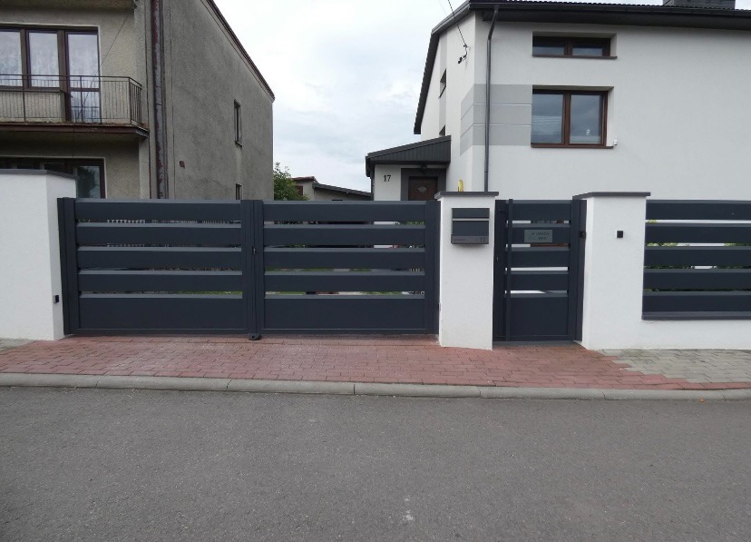 Nowoczesna, przesuwna brama w kolorze antracytowym z poziomymi panelami, osadzona na białych, murowanych słupkach. Widoczny fragment chodnika z kostki brukowej i elewacje budynków.