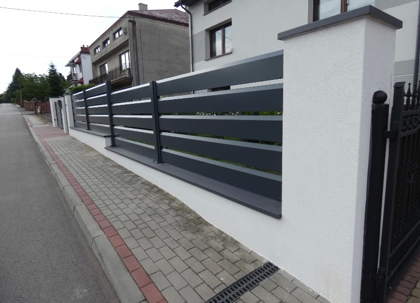 Nowoczesne ogrodzenie panelowe w kolorze grafitowym z białymi słupkami, widok z perspektywy ulicy, kostka brukowa i fragment bramy w tle.