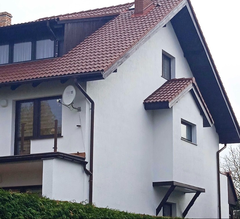 Biała elewacja domu jednorodzinnego z brązową dachówką i satelitą. Widoczne okna, rynny i fragment zieleni. Ujęcie z dołu, podkreślające bryłę budynku.