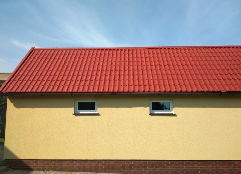 Żółta elewacja budynku z czerwonym dachem i dwoma oknami. Czerwona dachówka kontrastuje z kolorem elewacji. U dołu widoczny fragment muru z cegły.