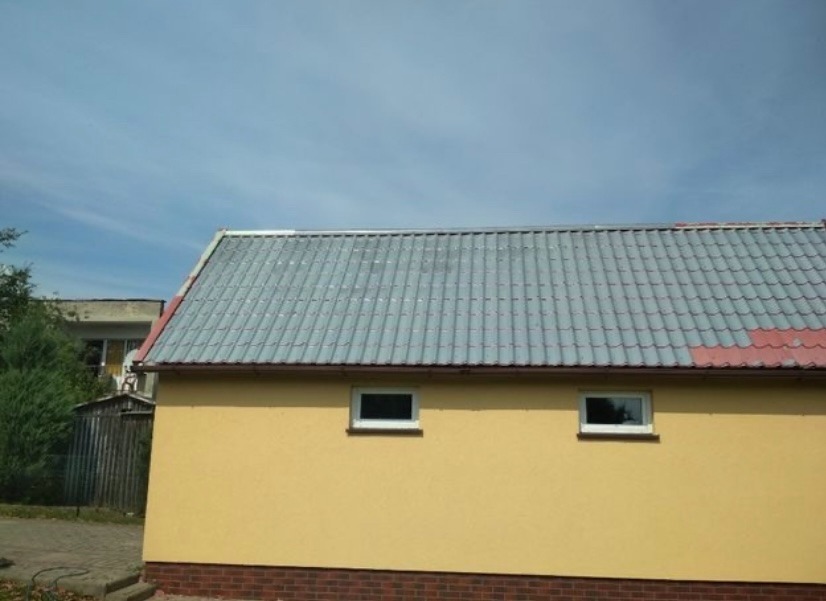 Żółty budynek z szarą dachówką i dwoma oknami, widok z dołu, fragment czerwonej dachówki w prawym górnym rogu, błękitne niebo w tle.