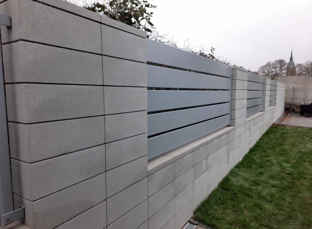 Nowoczesne ogrodzenie z szarego betonu i poziomych, metalowych przęseł, z zielenią w tle i fragmentem trawnika. Widok z perspektywy żabiej.