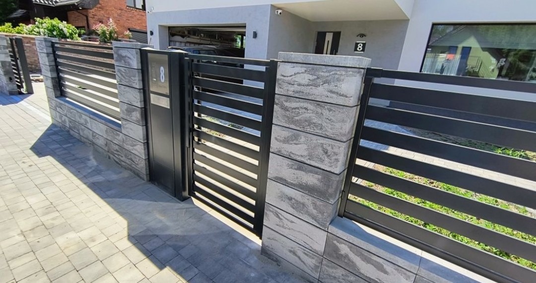 Nowoczesne ogrodzenie panelowe z bramką, szare betonowe słupki i poziome, czarne metalowe przęsła. Widok z zewnątrz posesji, kostka brukowa na podjeździe.