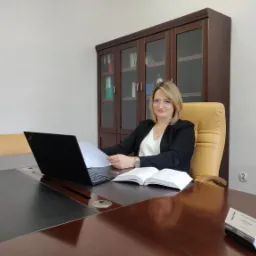 Kobieta w okularach siedzi przy biurku z laptopem i otwartą książką, w tle regał z dokumentami.