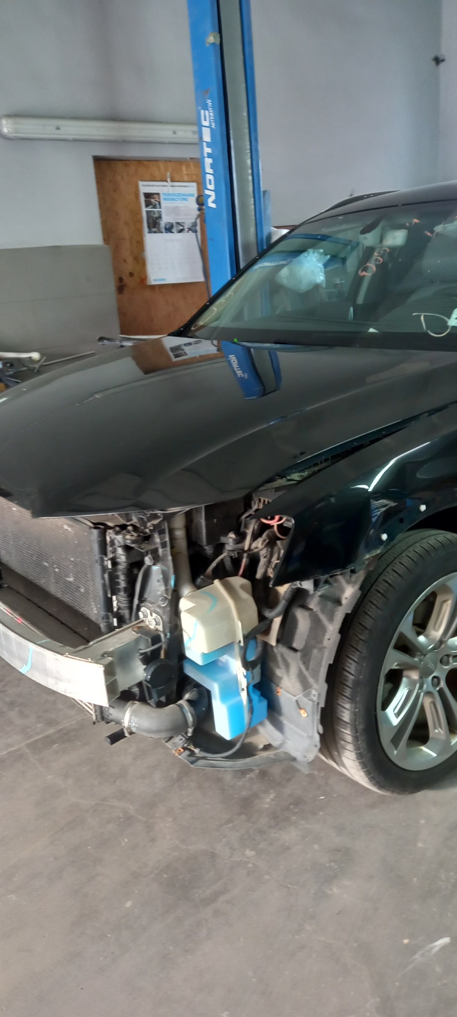 Uszkodzony przód czarnego samochodu na podnośniku w warsztacie, widoczny zbiornik płynu do spryskiwaczy i odsłonięta chłodnica.