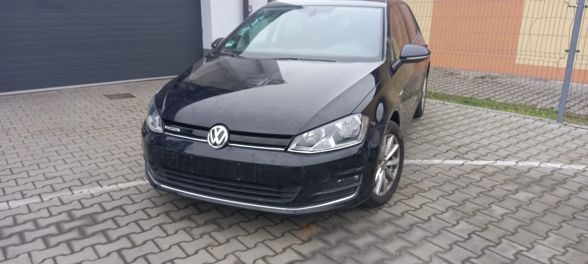 Czarny Volkswagen Golf Bluemotion zaparkowany na brukowanym podjeździe, z widocznymi detalami karoserii i felg.