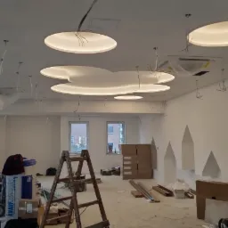 Wnętrze w trakcie montażu sufitu podwieszanego z okrągłymi elementami oświetleniowymi, widoczne kable, drabina, narzędzia i materiały budowlane na podłodze, w tle okna.