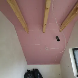 Poddasze w trakcie wykańczania: różowe płyty kartonowo-gipsowe zamontowane na drewnianych belkach, widoczne przewody instalacyjne i czarne worki na śmieci w rogu pomieszczenia.