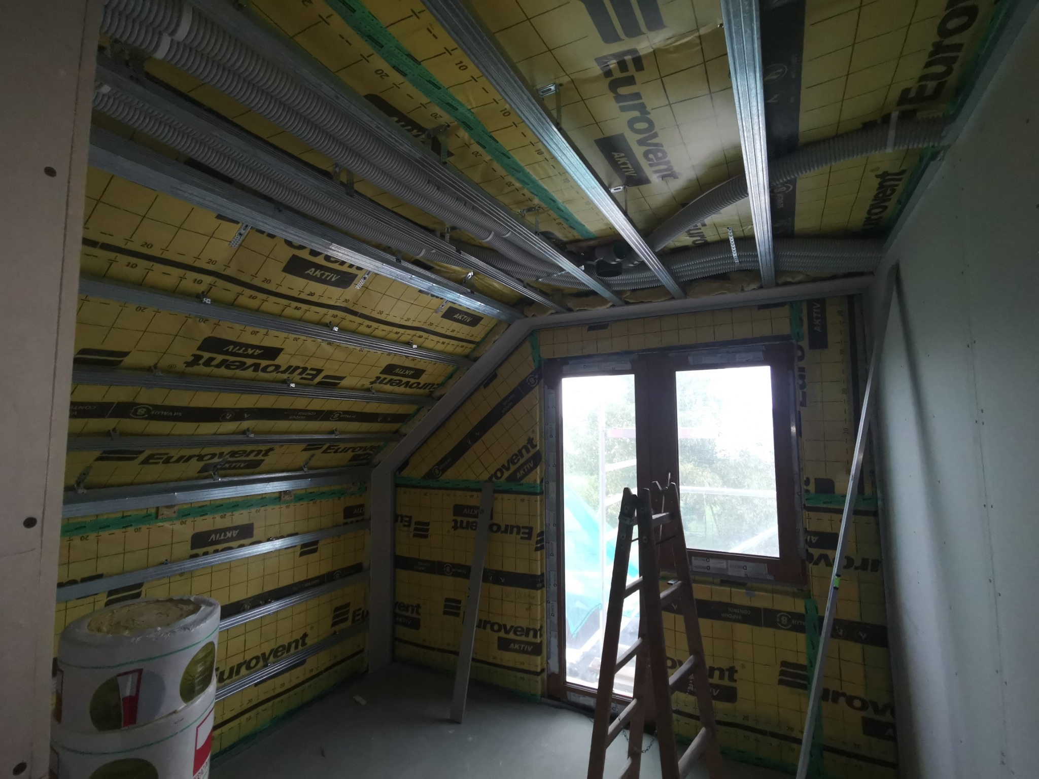 Poddasze w trakcie ocieplania, widoczne metalowe profile, izolacja Eurovent Aktiv, rury wentylacyjne i drabina oparta o okno.