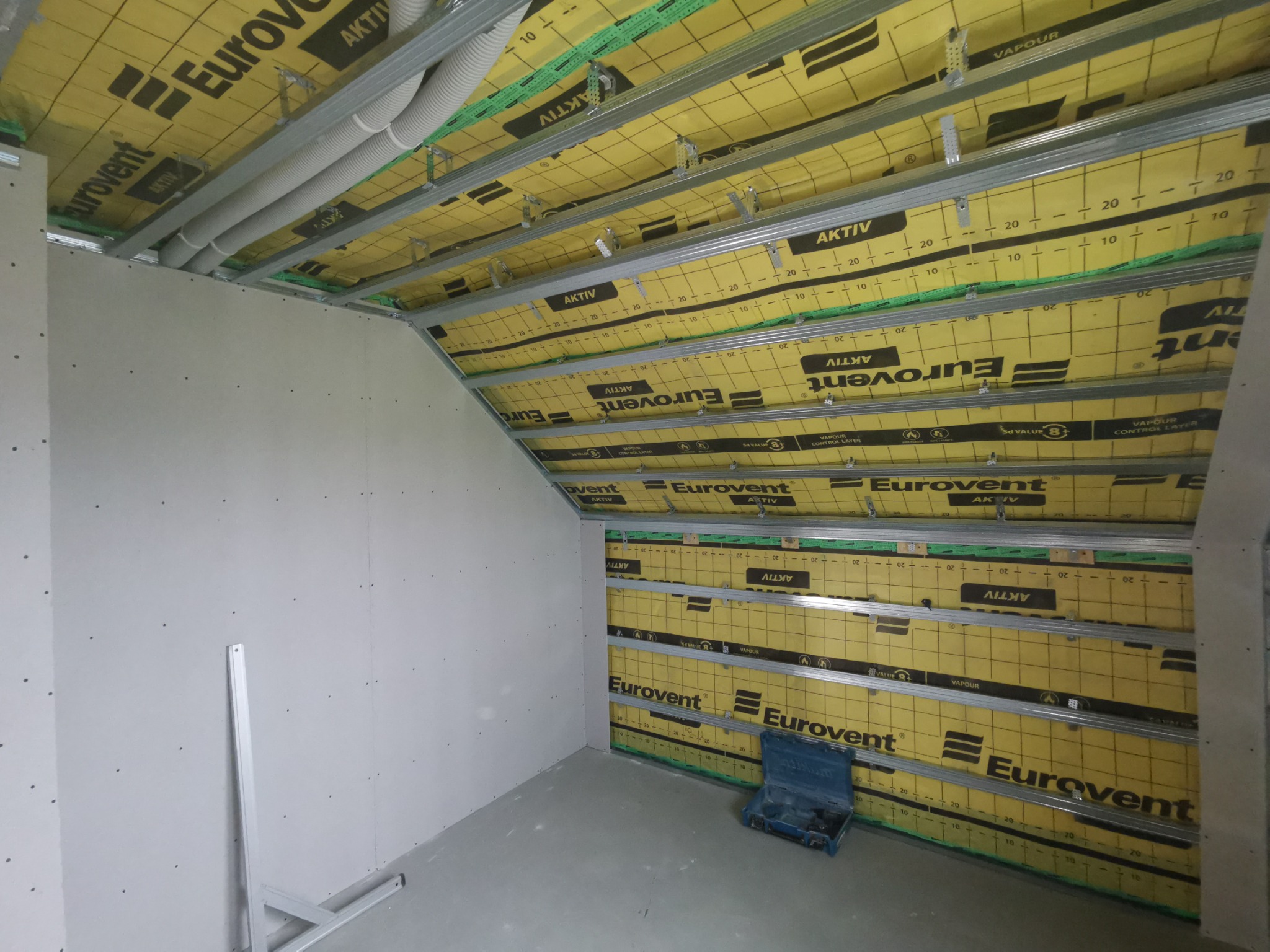 Poddasze w trakcie ocieplania z widoczną żółtą membraną Eurovent Aktiv, metalowym stelażem i częściowo zamontowanymi płytami karton-gips na ścianie szczytowej, z niebieską skrzynką narzędziową...