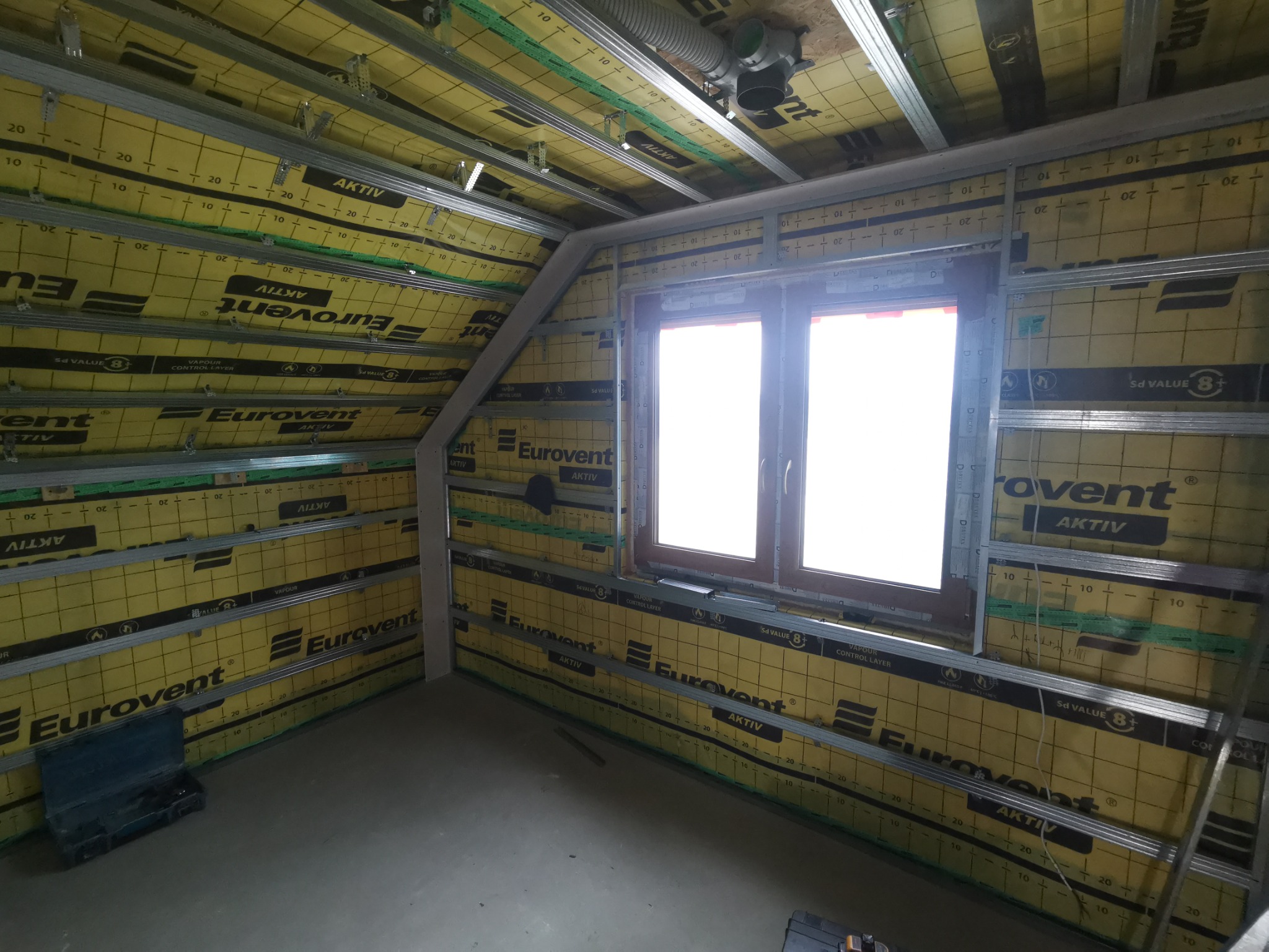 Poddasze w Biadolinach Szlacheckich w trakcie ocieplania, widoczne metalowe profile, żółta folia izolacyjna Eurovent z nadrukiem, okno oraz fragment instalacji wentylacyjnej pod sufitem.