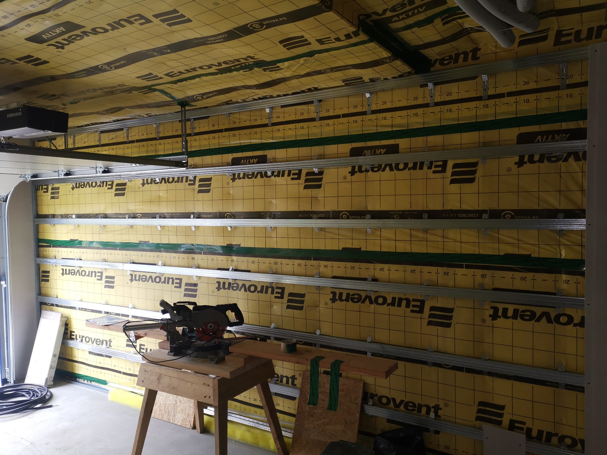 Poddasze w trakcie ocieplania z widoczną warstwą izolacji Eurovent, metalowym stelażem i narzędziami do cięcia na prowizorycznym stole warsztatowym.